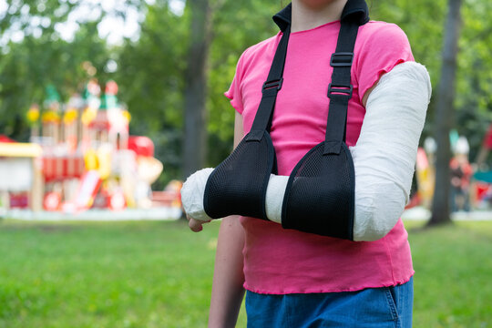 Girl with bone fracture in full arm cast and sling standing near playground summer. Childhood injury and safety concept