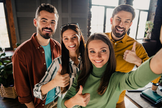 Four happy coworkers taking a selfie with thumbs up in a modern office, celebrating teamwork, friendship and success
