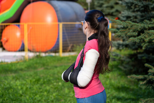 Girl with bone fracture in full arm cast and sling standing near inflatable trampoline. Childhood injury and safety concept