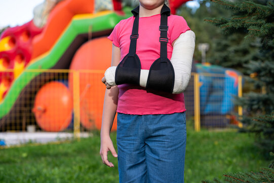 Girl with bone fracture in full arm cast and sling standing near inflatable trampoline. Childhood injury and safety concept