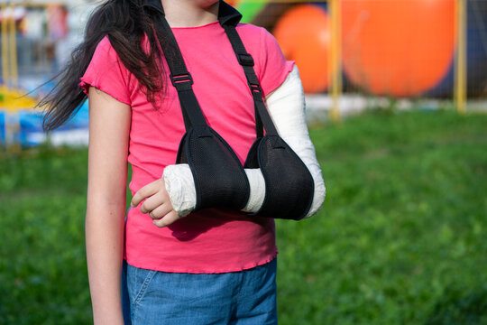 Girl with bone fracture in full arm cast and sling standing near inflatable trampoline. Childhood injury and safety concept