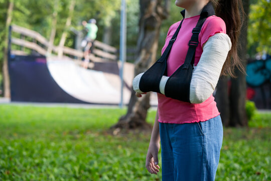 Girl with bone fracture in full arm cast and sling standing near skatepark summer. Childhood injury and safety concept