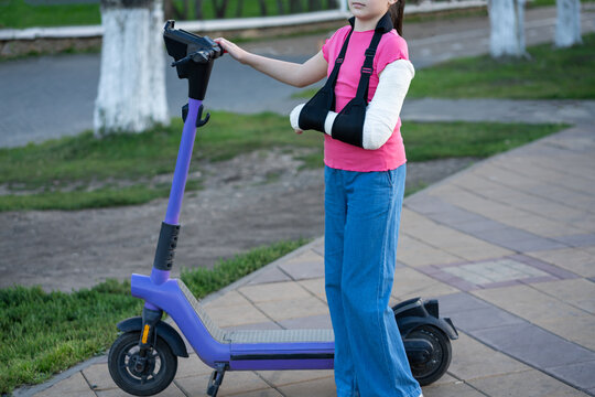 Unrecognizable girl with broken arm in plaster cast and sling standing near electric scooter, child injury and urban safety concept