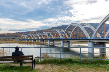 Man sits on a bench overlooking the railway arch bridge near Jezioro Mucharskie in an autumn mountain landscape. Scenic travel lifestyle concept of relaxation, solitude and peaceful leisure beside wat © Volha