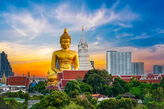 Large golden Buddha sits peacefully next to a white pagoda surrounded by lush trees and city buildings under a vibrant sunset sky in Bangkok