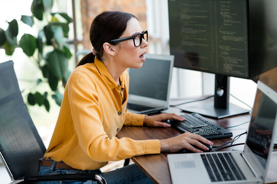 Surprised female programmer discovering error on multiple screens while coding in a bright modern office with laptops and a large monitor