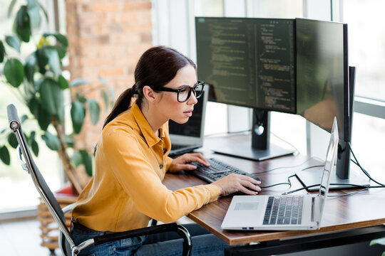 Female software developer concentrating on code while working at multiple monitors in a bright modern office with laptop and keyboard