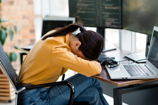 Exhausted programmer woman with head on desk after long coding session at office workstation with laptop and monitors &mdash; burnout and stress in tech workplace