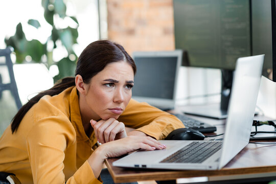 Frustrated young woman leaning on desk and staring at laptop screen in office, feeling tired and overwhelmed by work and computer tasks