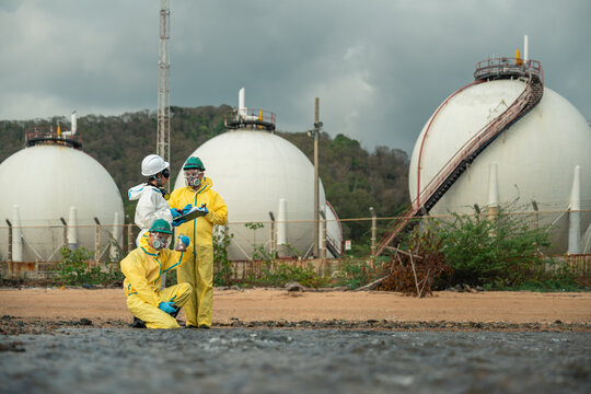 Environmental Workers Inspect Contaminated Water and Review Field Data Near Petrochemical Storage Tanks While Wearing Hazmat Suits, Respirators, Helmets, Goggles, and Gloves