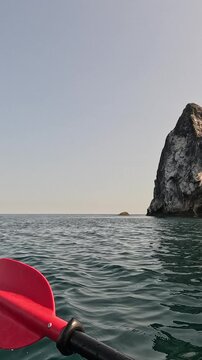 Red paddle dipping into calm ocean water beside a towering sea cliff. Handheld first-person shot follows the gentle movement of the vessel.