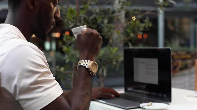 Man working on laptop. Black man working remotely on laptop and drinking coffee at outdoor cafe.