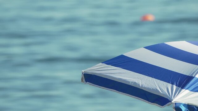Mediterranean sea waves ripple beneath a coastal sun umbrella as a distant buoy floats. Static extreme close-up shot.