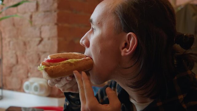 Close up of man holding and eating sandwich with visible ingredients like tomato lettuce. Background features brick wall potted plant on windowsill soft natural light creating warm indoor