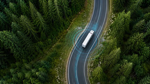 Aerial view of an autonomous truck driving along a winding highway through dense evergreen forest with digital sensor guidance illustrating connected logistics and intelligent transportation technolog