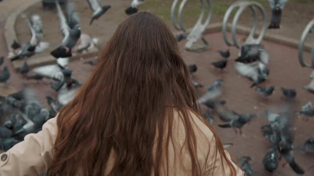 Close up of woman feeding pigeons in outdoor setting extending hand towards flock on ground. Pigeons gather around her feet and wings flap as woman interacts with them. Background includes