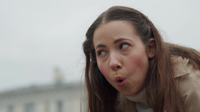 Close up of young woman with long brown hair wearing beige coat and white turtleneck showing expressive facial movements. Her hands gesture near her face against overcast urban background
