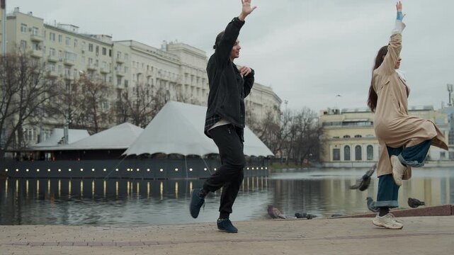 Close up of man and woman dancing together outdoors near lake with city buildings in background. Man wears dark jacket plaid shirt woman has long hair beige coat blue jeans. Both perform
