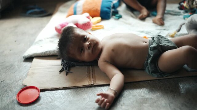 Portrait of a poor baby boy lying on a cardboard mat on the floor looking at the camera