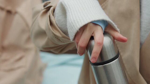 Close up of person wearing beige coat and white sweater unscrewing lid of stainless steel thermos. hand movement is steady as it opens container revealing its empty interior against soft