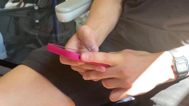 hands and forearms rest on lap while thumbs tap smartphone with vivid pink case, bright sunlight creates strong highlights, shadows, and casual travel mood inside