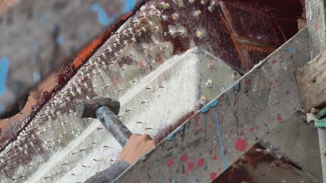 Worker hammering staples on fiberglass hull of traditional Vietnamese wooden fishing boat in shipyard, Central Vietnam