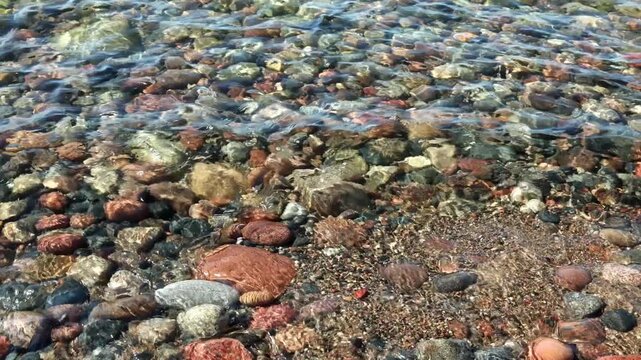 Clear sea water flowing over colorful pebbles on rocky shore in sunlight