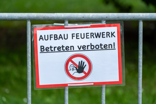 A red-bordered German sign reading fireworks setup, entry forbidden, hangs zip-tied to a metal barrier fence with greenery behind.