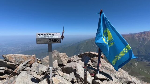 National flag of Kazakhstan fluttering next to 3680 meters altitude sign marker on top of Big Almaty Peak under clear blue sky