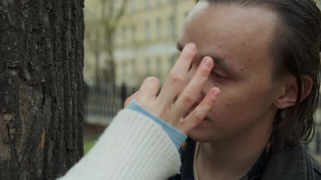 Close up of man standing near tree in outdoor urban setting looking around with minimal movement. Another man hand gently touches first subject on their face indicating interaction