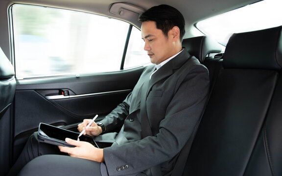 Asian businessman in suit using digital tablet with stylus pen on backseat of luxury car. Focused executive signing documents online, business mobility, remote work concept during vehicle transport