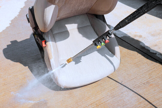 Man uses pressure washer to clean car seat in driveway water sprays onto fabric.