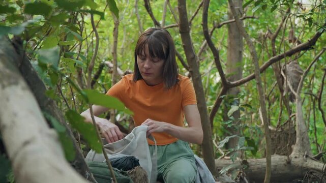 Woman in orange shirt and green pants sits among dense forest foliage interacting with bag of bread. Woman adjusts her position on ground surrounded by natural elements like tree trunks