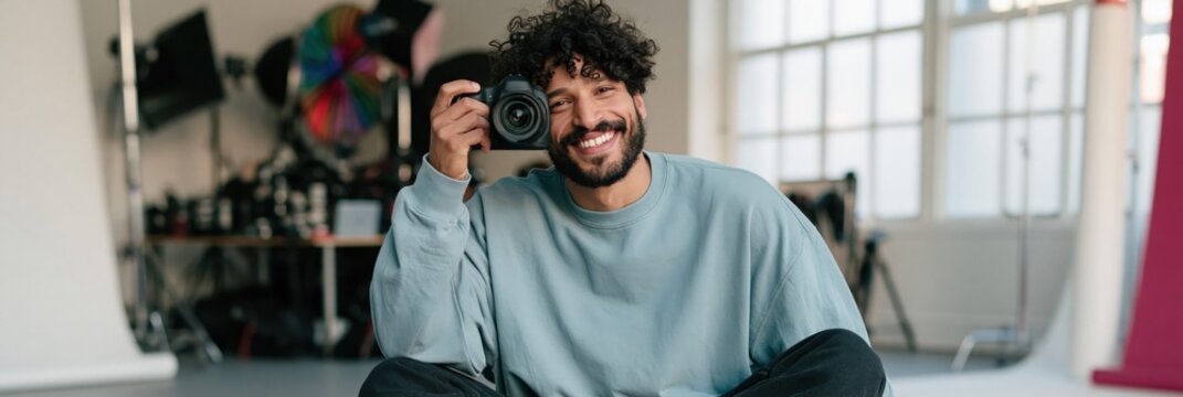 Smiling seated male photographer holding camera in wide studio scene for social media banner and website background
