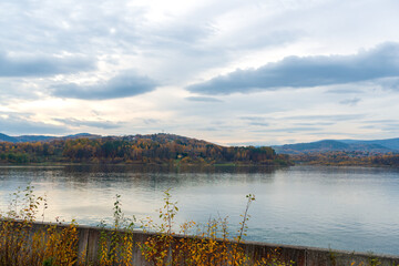 Wide view across Jezioro Mucharskie toward forested Beskid hills covered in golden and brown autumn foliage. Calm reservoir water and dramatic clouds create a scenic destination image for mountain tou © Volha