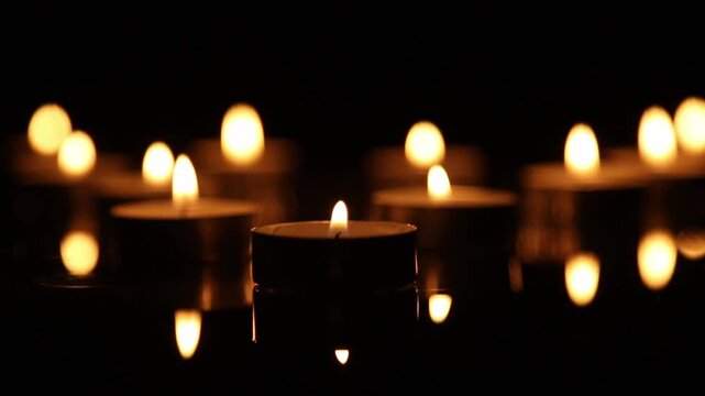 Multiple tealight candles glow warmly in a blurred row against a dark background, their soft flickering flames creating a golden bokeh effect with gentle reflections shimmering below.