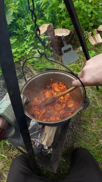 Man stirring frying carrots and onions in campfire cauldron for outdoor picnic cooking lifestyle