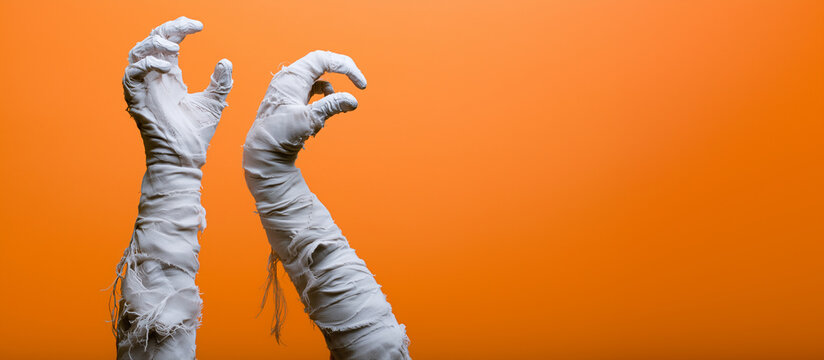 Two mummy hands wrapped in frayed white bandages reaching upward with curled fingers against a bright orange background, Halloween horror, undead arms, ancient curse, wrapped corps