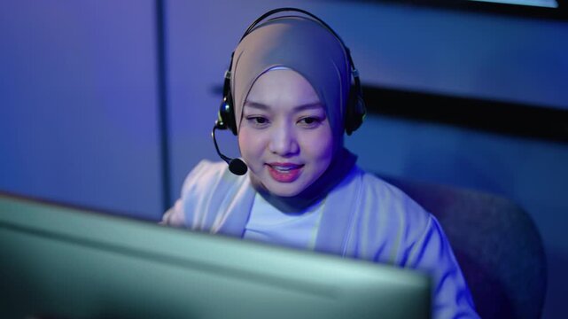 friendly Muslim woman wearing a hijab and headset works late at night in a call center office, providing online customer support, remote helpdesk assistance, and telemarketing client services.