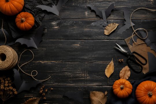 Halloween crafting flat lay with orange pumpkins, black paper bats, scissors, twine and dry autumn leaves on dark rustic wooden table.