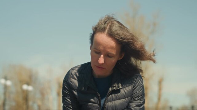 Close up of woman outdoors with wind blowing through her hair wearing dark puffer jacket and holding red pen. background features blurred trees under clear blue sky lighting emphasizing