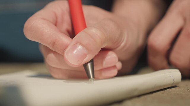 Close up of hand holding red pen writing on paper with soft lighting and focused detail. Hand moves slightly as it writes maintaining close proximity to white sheet throughout. Background