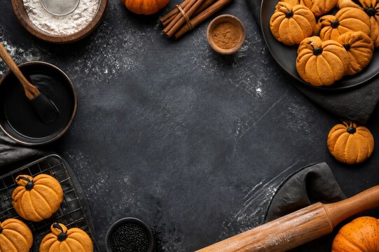 Halloween pumpkin cookies with baking ingredients on dark stone table. Flat lay with flour, cinnamon, chocolate glaze and rolling pin.