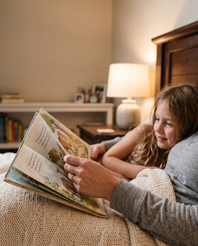 Adult reading storybook to child in cozy bedroom with warm lighting and soft blankets