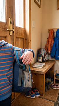 Child wearing striped hoodie holds backpack in home entryway with shoes and jackets nearby