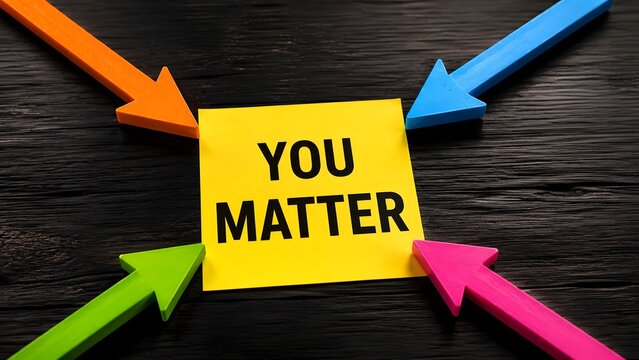 Yellow sticky note with text you matter on dark wooden desk surrounded by colorful pointing arrows for mental health awareness support and motivation
