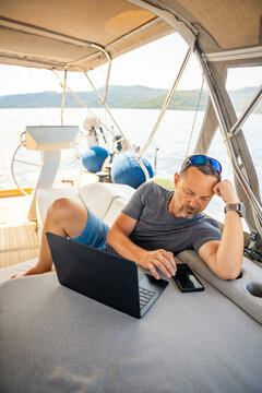 Relaxed man using laptop and smartphone while lounging on a sailing yacht. Digital nomad lifestyle and work life balance at sea.
