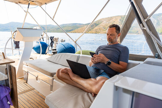 Man working remotely on a sailing yacht with laptop and smartphone. Location independent business and flexible lifestyle concept.