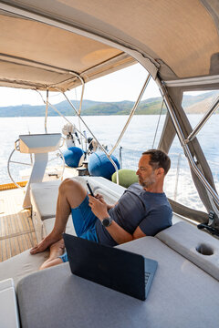 Man working remotely on a sailing yacht with laptop and smartphone. Location independent business and flexible lifestyle concept.