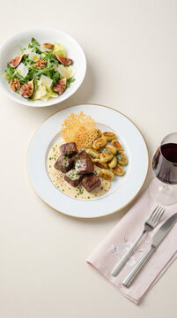 Grilled filet mignon cubes with handmade gnocchi, cheese sauce, crispy cheese, and a fresh salad with figs and walnuts, accompanied by a glass of red wine